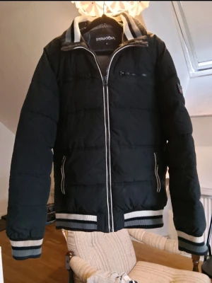 Svart pufferjacka med randiga detaljer - Svart pufferjacka från International med vita och grå randiga muddar vid ärmslut, krage och midja. Jackan har dragkedja framtill, två sidofickor med dragkedja och en bröstficka. Perfekt för kalla dagar och har en sportig vibe. L/XL