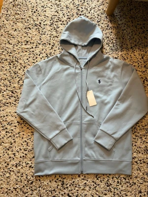 Ljusblå hoodie från Polo Ralph Lauren - Snygg ljusblå hoodie från Polo Ralph Lauren med dragkedja framtill och klassisk huva. Broderad logga på bröstet och två fickor. Perfekt för en chill och stilren look. Tillverkad i mjuk bomullsblandning.