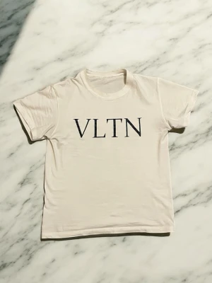 Vit VLTN t-shirt från Valentino - Vit t-shirt från Valentino med svart VLTN-tryck på bröstet. Klassisk rund halsringning och korta ärmar. Tillverkad i mjuk bomull för en skön och avslappnad känsla.