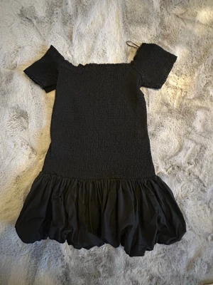 Populär  klänning från H&M - Supersöt svart kort klänning från H&M med smockad överdel och volangig kjol. Klänningen har korta ärmar och offshoulder-design som ger en trendig vibe. Perfekt för dig som gillar en enkel men snygg stil. Står tvärt inte vilken storlek det är men skulle gissa på S/M