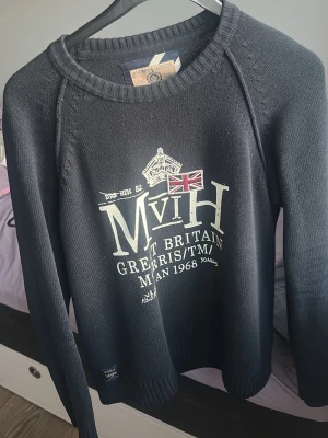 Mörkblå tröja från Morris Gents - Snygg mörkblå tröja från Morris med tryck framtill som inkluderar brittisk flagga och text. Tröjan har rund halsringning och ribbade muddar vid ärmslut och nederkant. Perfekt för en stilren och avslappnad look.