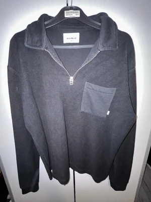 Svart halvzip-tröja från Woodbird - Svart långärmad tröja från Woodbird med halv dragkedja och krage. Tröjan har en bröstficka på vänster sida och är tillverkad i mjukt material. Perfekt för en chill och stilren look.