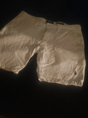 Vita linneshorts från Polo Ralph Lauren - Snygga vita shorts från Polo Ralph Lauren i linne. Klassisk passform med bälteshällor, knappgylf och två bakfickor med knappar. Liten broderad logga på ena benet. Perfekta för varma dagar.