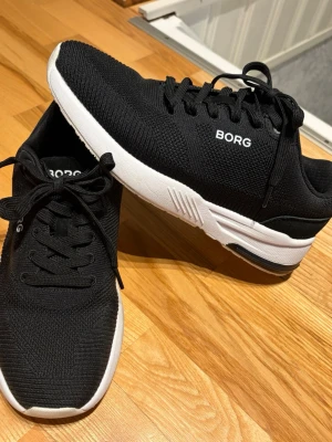 Svarta sneakers från BJÖRN BORG - Snygga svarta sneakers från BJÖRN BORG med vit sula och svart ovandel i meshmaterial. Skorna har klassisk snörning och låg profil, perfekt för en sportig och stilren look. Diskret logga på sidan och bekväm passform. Lättviktssko, endast använd en gång. Nyskick. 