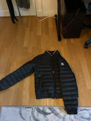 Svart pufferjacka från Moncler - Snygg svart pufferjacka från Moncler med quiltad design och diskret logga på bröstet. Jackan har dragkedja framtill, två sidofickor med dragkedja och en krage med rödvit detalj. Perfekt för vår, höst och vanliga sommar dagar. riktigt stilren look.