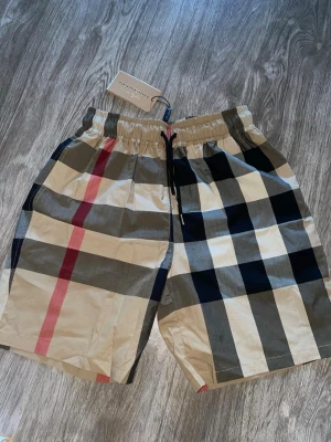 Rutig Burberry shorts med snörning - Säljer ett par snygga Burberry shorts med klassiska Nova Check-rutorna. Storlek M finns på dessa, men finns även i S och L. Elastisk midja med snörning för bra passform. Lätta och sköna – perfekta till sommaren, stranden eller vardagsbruk.  * Märke: Burberry * Storlek: M (finns även S & L) * Skick: Mycket fint skick / oanvända med tags kvar * Färg: Beige/rutig * Äkta detaljer och märkning finns på bilderna  Skickas snabbt 📦 Skriv vid frågor eller för fler bilder!