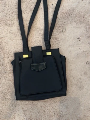 Svart fyrkantig axelväska med ficka - Stilren svart axelväska i fyrkantig form med lång axelrem. Väskan har en stor ficka framtill med lock och dragkedja samt gulddetaljer vid handtagen. Materialet ser ut att vara syntetiskt, vilket ger en modern och clean look.