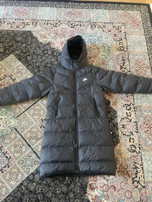 Svart lång dunjacka från Nike - dags för mig att sälja min svarta långa dunjacka från Nike med huva och dragkedja framtill. Jackan har två praktiska fickor med dragkedja och diskret Nike-logga på bröstet. Perfekt för kalla vinterdagar med sin vadderade design och stilrena look.