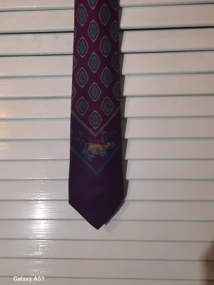 Lila slips från Boss med mönster - Säljer en lila slips från Boss med färgstarkt geometriskt mönster i blått och rött samt ett motiv av en räv i naturmiljö. Slipsen är tillverkad i 100% siden som känns lyxigt och sticker ut med sin unika design.