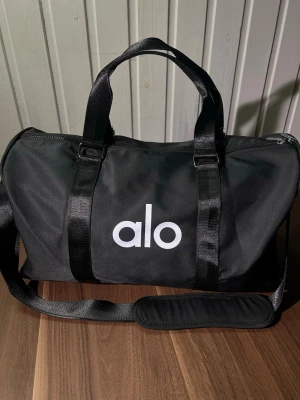 Svart resväska från Alo - Snygg svart resväska från Alo med vitt logotryck på framsidan. Väskan har både bärhandtag och justerbar axelrem för smidig transport. Tillverkad i slitstarkt tygmaterial och har dragkedja upptill. Perfekt för weekendresor eller gymmet.