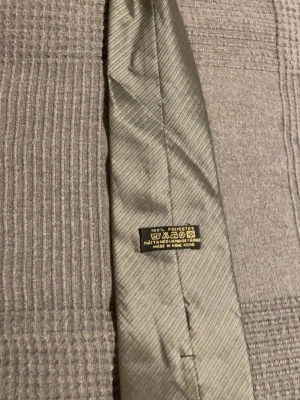 Ljusgrå slips i polyester - Snygg ljusgrå slips med diskret randigt mönster. Tillverkad i 100% polyester och har en klassisk form. Perfekt accessoar för att lyfta din outfit med en stilren touch.