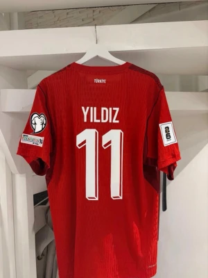 Turkiet fotbollströja #Yildiz - Röd fotbollströja från Nike med Turkiets flagga på bröstet och nummer 11 samt namnet Yildiz på ryggen. Tröjan har korta ärmar, vita detaljer och tryck på båda ärmarna. Snyggt mönster i tyget och sportig passform. Matchtröjan är i player fit så den är slim model och därför motsvarar ungefär Xl/L