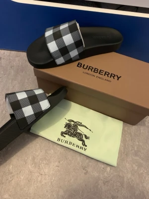 Burberry rutiga svarta tofflor - Snygga tofflor från Burberry med klassiskt svartvitt rutmönster på remmen. Mjuk och bekväm sula i svart syntetmaterial. Perfekta för chill dagar eller när du vill ha en stilren look. Levereras med originalkartong och dustbag.