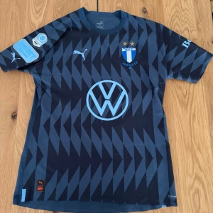Malmö FF matchanvänd tröja blå - Snygg mörkblå Malmö FF matchtröja från Puma med ljusblå detaljer och tryck. Tröjan har klubbmärke, sponsorer som Volkswagen och Unibet samt Allsvenskan-märke på ärmen. Tillverkad i lätt och ventilerande material, perfekt för fotbollsträning eller match. Vunnen på Malmö ff egen auktion 