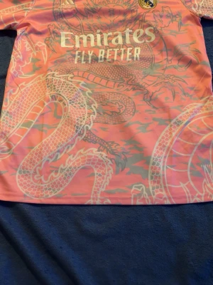 Rosa  Real Madrid tröja - Säljer en rosa Real Madrid t-shirt från Adidas med stort drakmönster i vitt och grått. Den har knappt använts och köptes förra sommaren, Storleken är L men passar också M. Kom priv för fler frågor