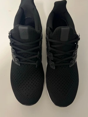 Adidas Ultra Boost svarta sneakers - Svarta Adidas Ultra Boost sneakers med stickad ovandel i mesh och klassiska tre ränder på sidan. Skorna har svart snörning och vit Boost-sula för extra komfort. Perfekta för en sportig och stilren look.