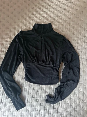 Svart glittrig polotopp med lång ärm - Snygg svart glittrig polotopp med lång ärm och lätt croppad passform. Toppen har en liten slits framtill och är tillverkad i ett stretchigt material som ger en bekväm känsla. Storleken är M men har även passat mig som normaltsätt är S på grund av det stretchiga materialet 