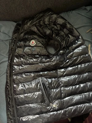  Moncler Väst - Snygg svart dunväst från Moncler med glansig finish och klassisk logga på bröstet. Västen har dragkedja framtill och en praktisk ficka med dragkedja. Perfekt för lager-på-lager och streetstyle. Materialet är lätt och isolerande.
