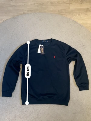 Mörkblå tröja från Polo Ralph Lauren - Snygg mörkblå tröja från Polo Ralph Lauren med klassisk rund hals och röd broderad logga på bröstet. Tröjan har långa ärmar, ribbade muddar och är tillverkad i mjukt material. Perfekt för en stilren och avslappnad look. Helt ny och aldrig använd. Är aktiv 24/7 svarar direkt✅ Fraktar antigen samma dag eller nästa. Är väldigt snabb på att lämna paket o sånt🚚📦Hör av dig via frågor⁉️