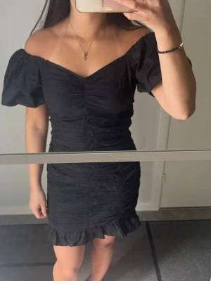 Svart offshoulder kort klänning - Trendig svart kort klänning med offshoulder och puffärmar. Klänningen har rynkade detaljer längs hela kroppen och en volangkant nertill. Perfekt för dig som gillar en feminin och modern stil.
