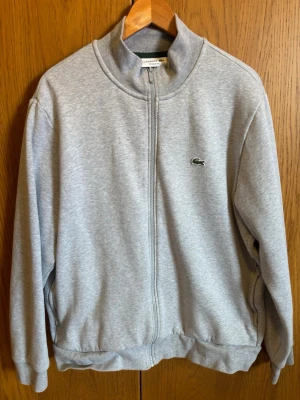 Grå ziptröja från Lacoste - Grå ziptröja från Lacoste med hög krage och dragkedja framtill. Den är i storlek XXL men passar även XL. Tröjan har den klassiska gröna krokodilloggan broderad på bröstet och ribbade muddar vid ärmslut och nederkant. Tillverkad i mjuk bomullsblandning för en chill och clean look.