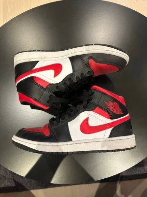 Air Jordan 1 Mid svart/röd/vit - Snygga Air Jordan 1 Mid sneakers i svart, rött och vitt läder. Klassisk design med röd swoosh, svart snörning och ikoniska Jordan Wings-loggan på sidan. Perfekta för dig som gillar streetstyle och vill sticka ut med en tidlös modell.