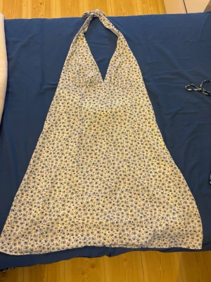 Vit och blå blommig halterneckklänning - Söt somrig klänning med halterneck och v-ringning. Vit botten med små blå blommor över hela klänningen. Lätt utsvängd modell som knyts i nacken. Perfekt för varma dagar och har en luftig känsla.