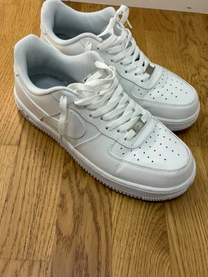 Nike Air Force 1 vita sneakers - Klassiska Nike Air Force 1 sneakers i helvitt skinn med perforerad tå, snörning och ikonisk swoosh på sidan. Tjock sula och bekväm passform, perfekt för dig som gillar stilrena och tidlösa sneakers.