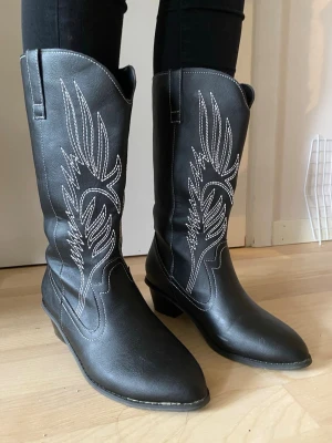 Svarta cowboy boots med vit brodyr - Snygga svarta cowboy boots med spetsig tå och vit dekorativ brodyr längs skaftet. Klassisk westernstil med medelhöga klackar och dragflikar upptill. Tillverkade i konstläder med coolt mönster som ger bootsen en unik look. Använda en gång så de är i fint skick!☺️