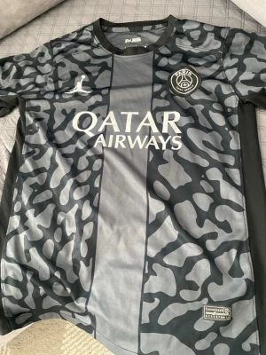 PSG x Jordan matchtröja svart/grå - Snygg PSG matchtröja i samarbete med Jordan Brand. Tröjan är svart och grå med ett unikt kamouflagemönster, PSG-logga på bröstet och Qatar Airways som sponsor. Tillverkad i lätt och ventilerande funktionsmaterial, perfekt för fotboll eller träning.
