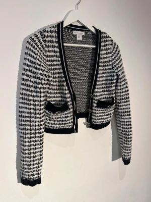 Svartvit kofta från London Kaye - Snygg svartvit kofta från London Kaye med stickat mönster, svarta kanter och två fickor framtill. Kortare modell med dekorativa spännen framtill. Perfekt för att lyfta din outfit med en trendig touch.