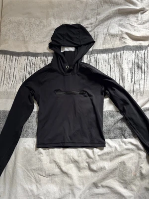 Svart croppad tränings hoodie - Svart croppad hoodie i stretchigt syntetmaterial med lång ärm och huva. Toppen har en dragkedja framtill som detalj och ger en sportig look, perfekt för träning eller streetwear.