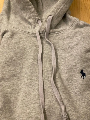 Grå hoodie från Polo Ralph Lauren - Snygg grå hoodie från Polo Ralph Lauren med klassisk broderad logga på bröstet. Hoodien har huva med dragsko och en stor magficka. Tillverkad i mjukt bomullsmaterial och har ribbade muddar vid ärmslut och nederkant.