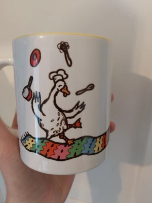 Mugg med tecknad hjälpsam anka - Vit keramikmugg med färgglad illustration av en tecknad anka med kockmössa som jonglerar köksredskap. Texten 'Hjälpande and' och ett mönster i regnbågsfärger runt nederkanten. Perfekt för varma drycker.