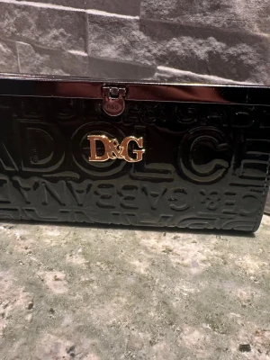 Svart plånbok med gulfärgat emblem - Snygg svart plånbok med glansig finish.  Plånboken har ett guldfärgat emblem på framsidan och metallspänne upptill. Perfekt för dig som gillar lyxiga accessoarer med attityd. Köpt i Spanien. 