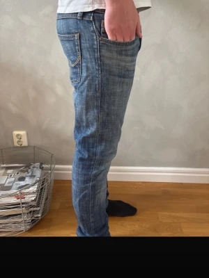 Blå slim fit jeans med slitningar - Säljer ett par blå slim fit jeans med snygga slitningar och ljusare partier på låren. Jeansen har klassisk femficksdesign och knappgylf. Perfekta för en avslappnad och trendig look.