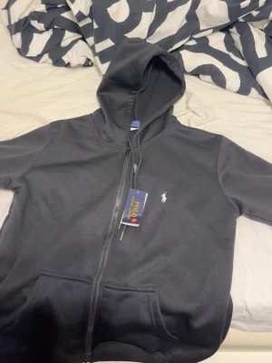 Svart hoodie från Polo Ralph Lauren - Svart hoodie med dragkedja från Polo Ralph Lauren. Klassisk design med huva och dragsko, två fickor framtill och den ikoniska vita logotypen broderad på bröstet. Perfekt för en chill och stilren look.