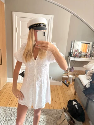 Vit kort klänning med spetsdetaljer/ studentklänning  - Superfin vit kort klänning med v-ringning, puffärmar och spetsdetaljer. Klänningen har knappar framtill och är perfekt för student eller sommar. Söt och feminin design med broderade mönster och lätt, luftig känsla.