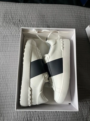 Valentino Garavani vita sneakers - Snygga vita sneakers från Valentino Garavani med en bred svart rand över mitten. Skorna är i läder och har coola nitar på sulan vid hälen. Perfekta för dig som vill ha en stilren men ändå unik look.