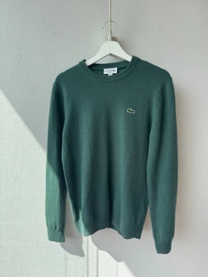 Grön stickad tröja från Lacoste - Mörkgrön stickad tröja från Lacoste med rund halsringning och klassisk krokodillogga på bröstet. Tröjan har långa ärmar och ribbade muddar vid ärmslut och nederkant. Perfekt för lager-på-lager och enkel att matcha. Använd 1-2 gånger (9.75/10)