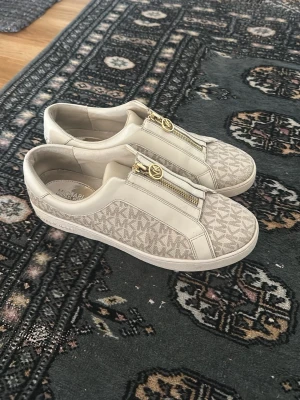 Beige Michael Kors sneakers med dragkedja - Skorna är ej så slitna som dom ser ut på bild, de defekter som syns är sånt som skapats utav dragkedjans metall. Jag ska såklart försöka att återgärda dessa defekter så att de blir mindre synliga. Priset kan diskuteras!