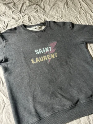 Grå Saint Laurent sweatshirt - Säljer en grå sweatshirt från Saint Laurent med coolt tryck i pastellfärger och blixt på bröstet. Tröjan har rund halsringning, ribbade muddar och är tillverkad i mjukt bomullsmaterial. Perfekt för en avslappnad och trendig look.