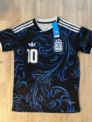 Argentina Messi VM-2026 Fotbollströja - Snygg svart och blå Argentina fotbollströja med Messi och nummer 10 på ryggen. Tröjan har vita detaljer, AFA-logga och Adidas-logga framtill samt coolt blått mönster över hela. Perfekt för fotbollsfans! HELT NY