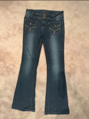Blå bootcut jeans med fickdetaljer - Säljer ett par blå bootcut jeans med snygga fickdetaljer och kontrastsömmar. Jeansen har dubbla knappar i midjan och dekorativa ficklock fram. Perfekta för dig som gillar en klassisk men ändå trendig look.
