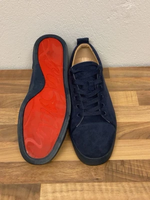 Mörkblå Christian Louboutin sneakers - Snygga mörkblå sneakers från Christian Louboutin i mocka med klassisk röd sula och diskret logga undertill. Skorna har rund tå, platta skosnören och låg profil. Perfekta för dig som vill sticka ut med exklusiv stil.