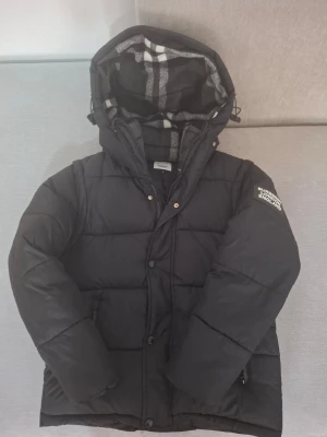 Svart pufferjacka Burberry England - Svart pufferjacka från Burberry med huva och rutigt innerfoder. Jackan har dragkedja och knappar framtill samt logga på ärmen. Klassisk quiltad design och varm vaddering, perfekt för kalla dagar.