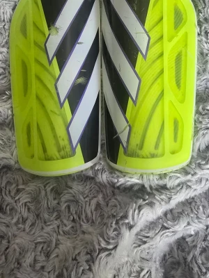 Adidas benskydd i neon och svart - Säljer ett par Adidas benskydd i neon-gult och svart med vita detaljer. De har en sportig design med tydliga ränder och är tillverkade i plast och skumgummi för bra skydd och komfort. Perfekta för fotbollsträning eller match.