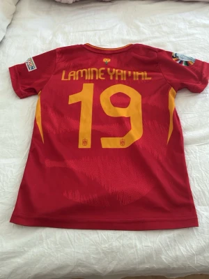 Spansk landslagströja Lamine Yamal - Röd matchtröja från Adidas för spanska landslaget med Lamine Yamal och nummer 19 i gult på ryggen. Tröjan har tryckta detaljer på ärmarna och är tillverkad i lätt, ventilerande polyester. Perfekt för fotbollsträning eller att bära som supporter.