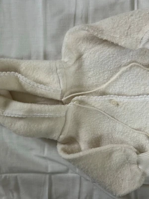 Vit teddyjacka med huva - Mysig vit teddyjacka med huva och knäppning framtill. Jackan har ribbade muddar vid ärmsluten och är tillverkad i ett fluffigt, mjukt material som ger en skön och avslappnad look.