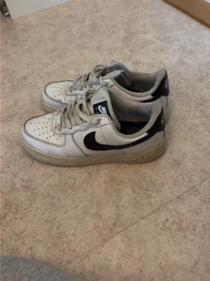 Nike Air Force 1 vita och svarta sneakers - Nike Air Force 1 sneakers i vitt läder med svarta detaljer och klassisk swoosh-logga på sidorna. Skorna har perforerad tå, platt sula och snörning framtill. Perfekt för dig som gillar streetwear och tidlös stil.